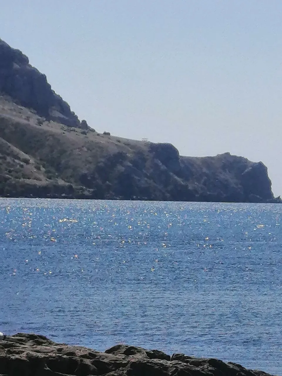 Скалистый мыс над морем и солнечные блики на воде, каменистый берег на переднем плане